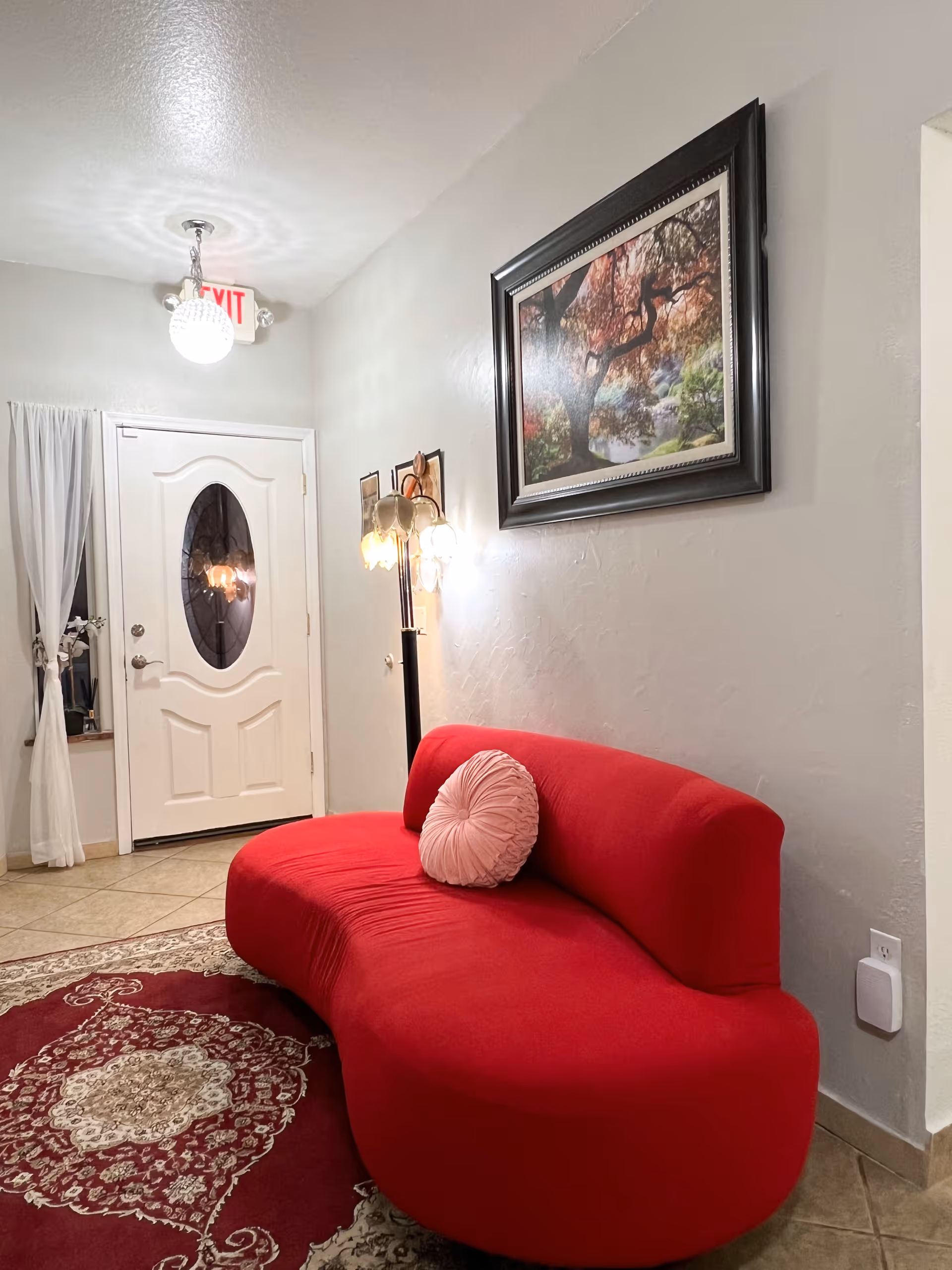 Interior view of a room with a red curved sofa featuring a round pink pillow, a patterned red and beige rug, a standing lamp with multiple bulbs, a framed painting of a tree with autumn leaves on the wall, a white door with an oval glass window, and a window with sheer white curtains.