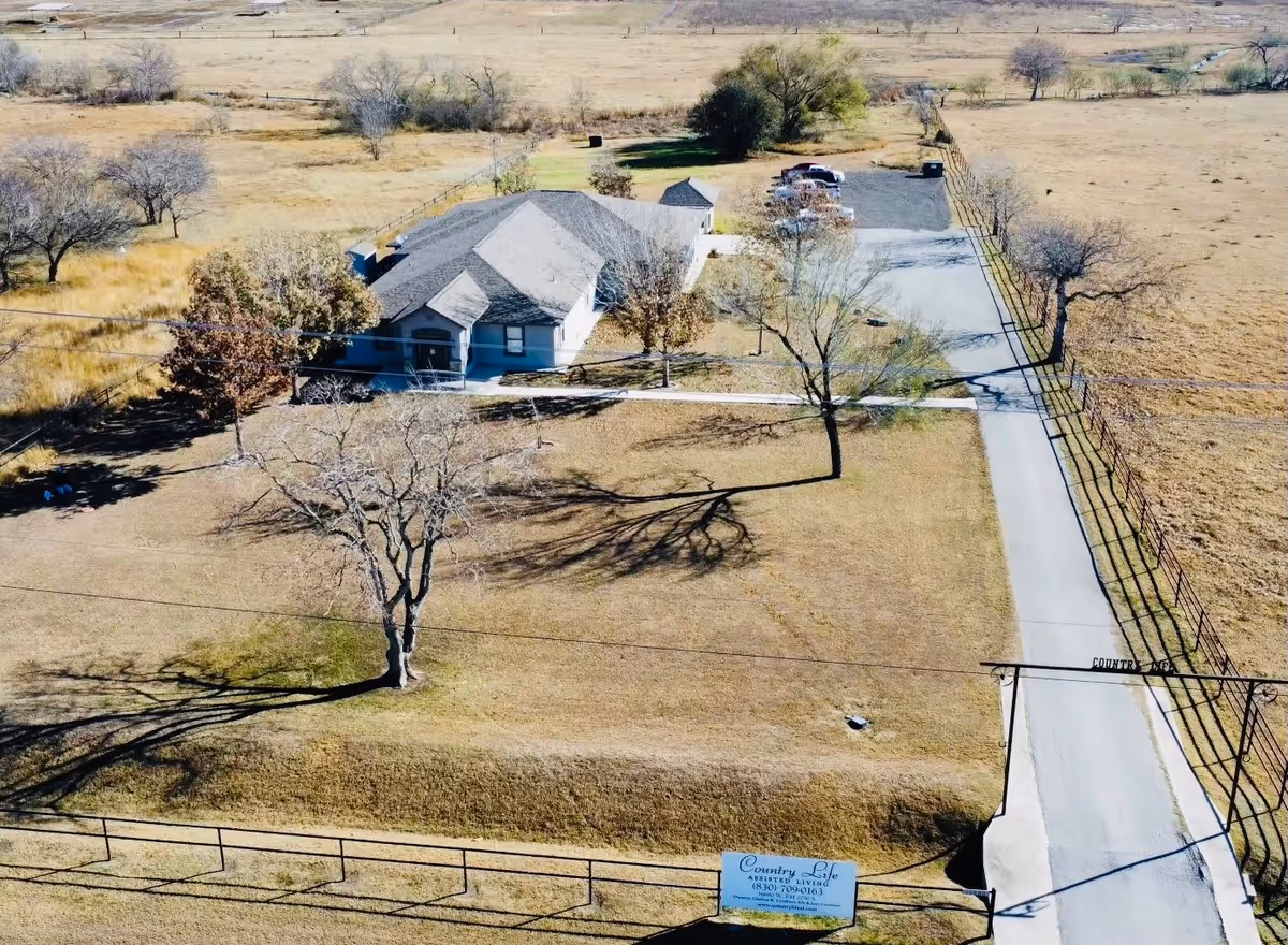 Aerial view of Country Life Assisted Living facility showing a single-story building with a gray roof surrounded by a large fenced grassy area with several leafless trees. A paved driveway leads to a small parking area with several cars. A sign with the facility's name and contact information is visible near the entrance.