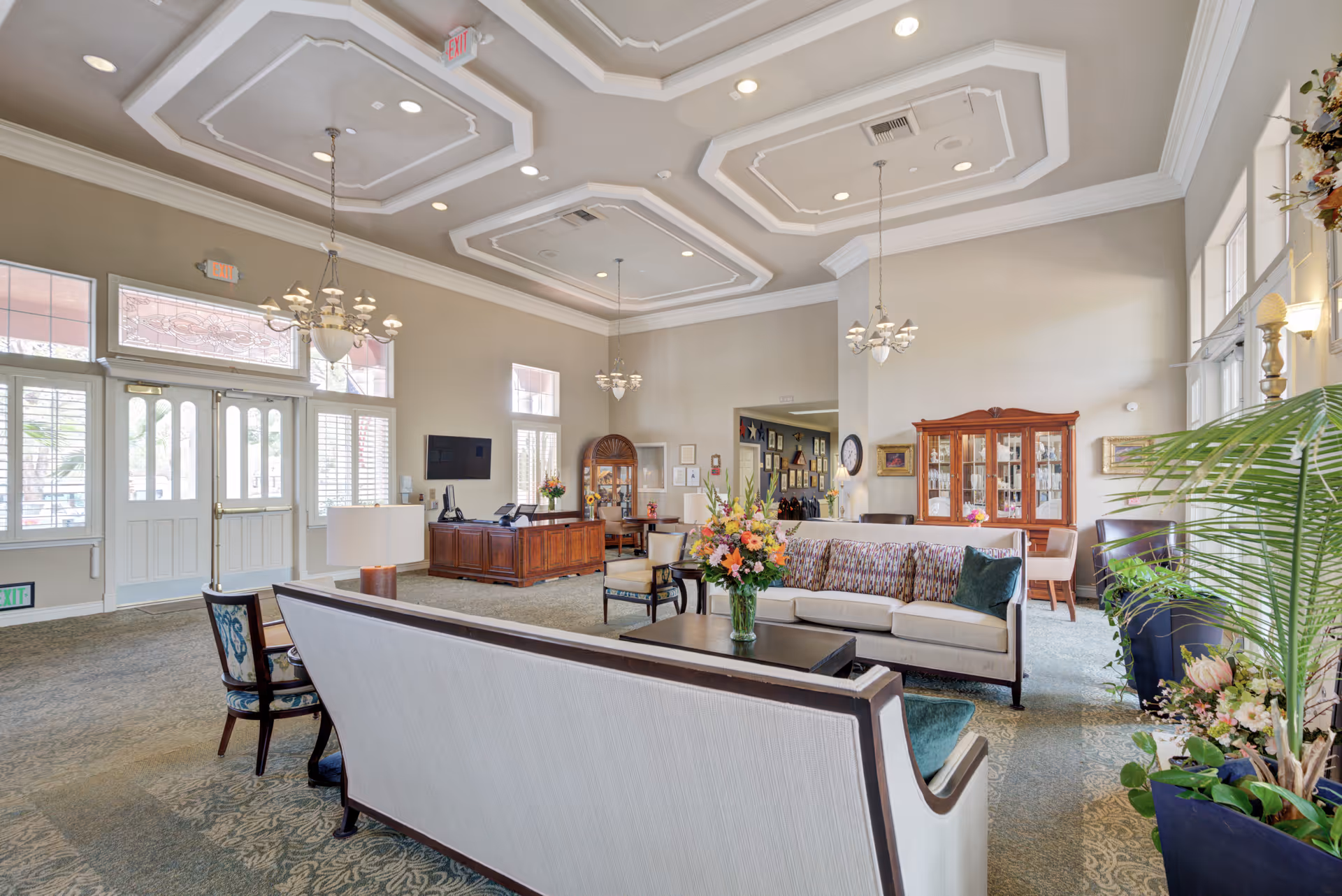 A spacious and elegant living room area in a senior living facility with high ceilings, chandeliers, and large windows allowing natural light. The room features comfortable sofas and chairs arranged around a coffee table with a vase of flowers. There is a wooden reception desk with a computer and a TV mounted on the wall. Decorative plants and a wooden cabinet with glass doors are also visible.