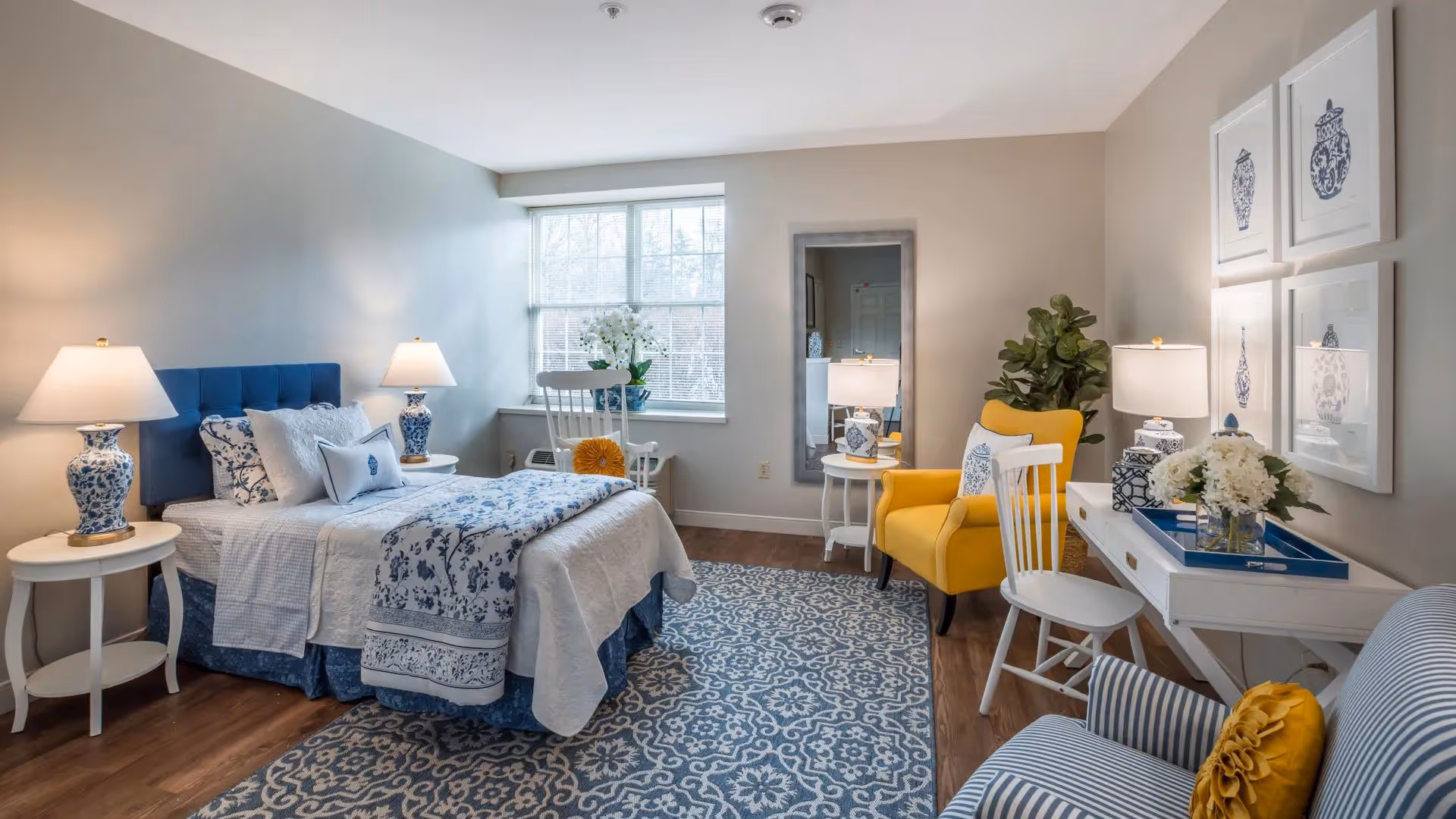 A cozy bedroom with a blue and white color scheme featuring a bed with floral bedding, two bedside tables with matching lamps, a large window, a yellow armchair, a white desk with a chair, decorative framed artwork on the wall, a patterned rug, and a striped armchair with a yellow pillow.