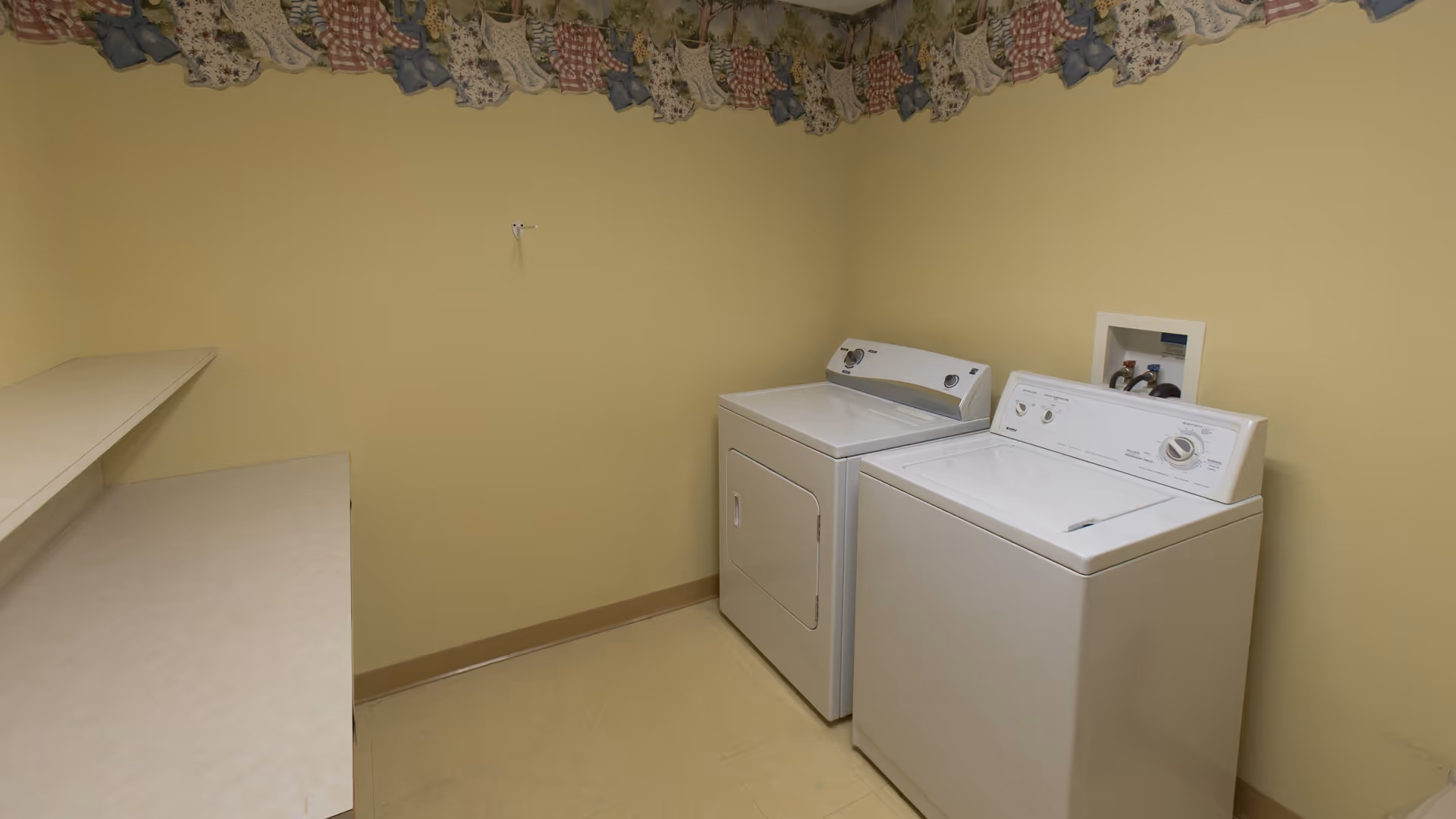 A laundry room with a white washing machine and dryer side by side against a yellow wall. There are two beige countertops on the left side and a decorative border with a floral and checkered pattern near the ceiling.