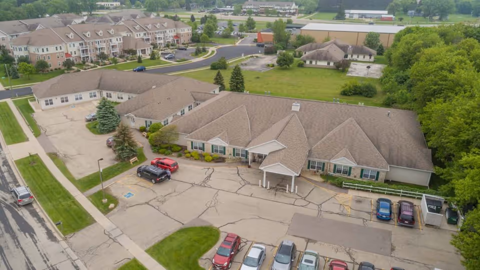 Aerial view of Stoughton Meadows Assisted Living facility showing a single-story building with a pitched roof, surrounded by parking spaces with several cars parked. The building is adjacent to a larger multi-story residential complex and is bordered by green lawns and trees.