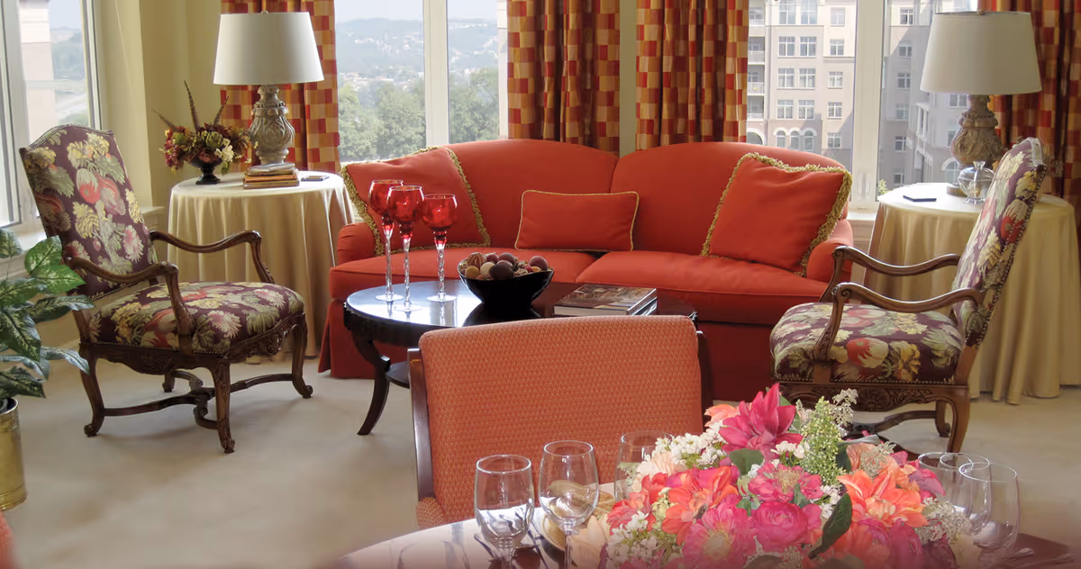 Sunlit living room with a red sofa, patterned armchairs, a round coffee table and a floral centerpiece by large windows.