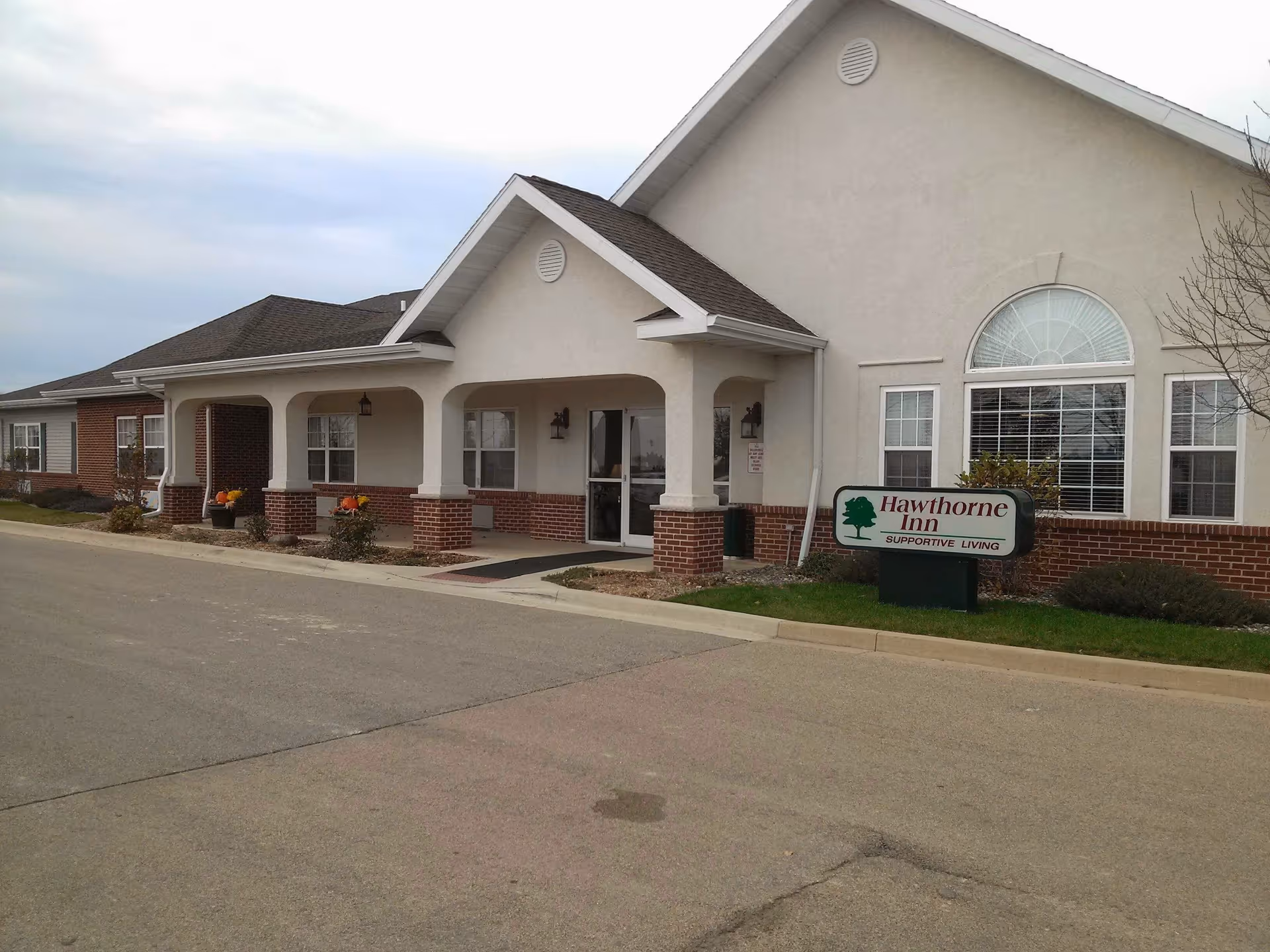 Exterior view of a single-story supportive living facility named Hawthorne Inn, featuring a covered entrance with white columns and brick accents, large windows, and a sign on the lawn.