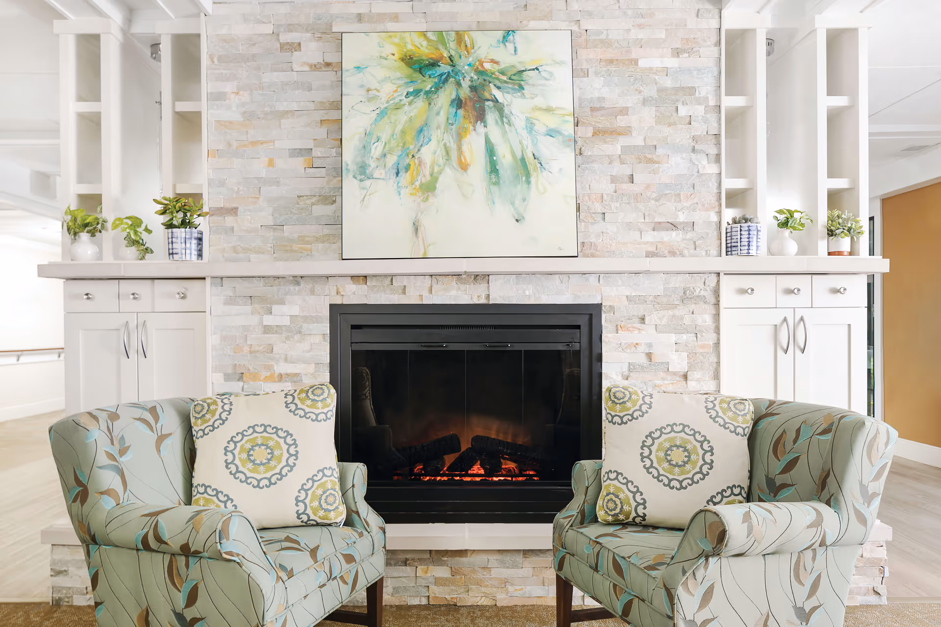 Two patterned armchairs with decorative pillows are positioned facing each other in front of a modern stone fireplace with a glowing fire. Above the fireplace hangs a colorful abstract painting. White built-in cabinets with small plants on top flank the fireplace on both sides.