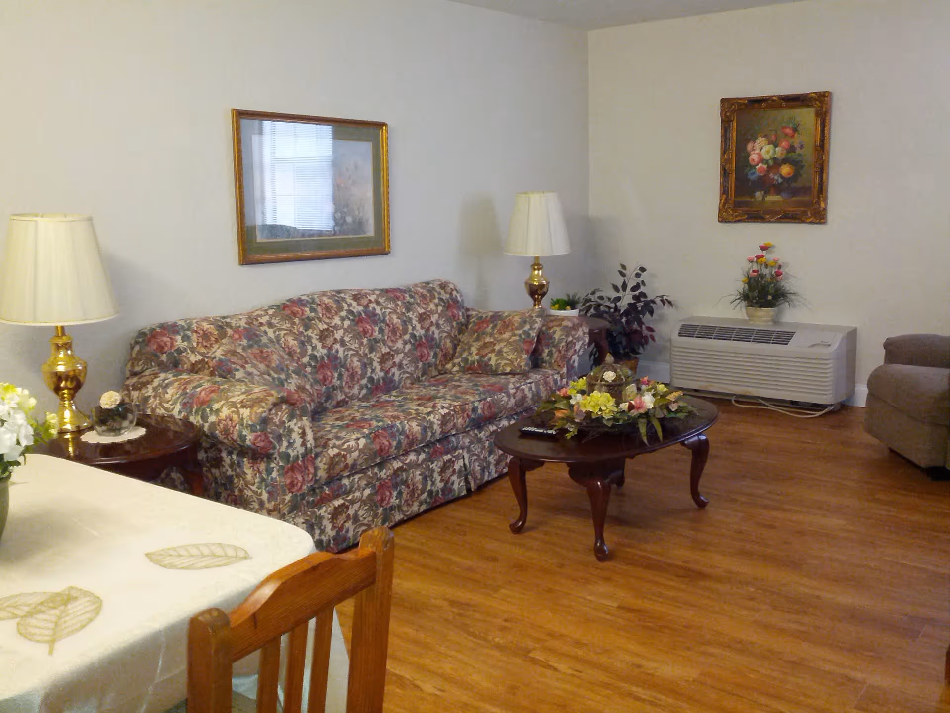 A cozy living room with a floral patterned sofa, two table lamps with white shades on wooden side tables, a wooden coffee table with a floral centerpiece, a beige armchair, and framed floral paintings on the walls. The room has wooden flooring and a white wall-mounted air conditioning unit.