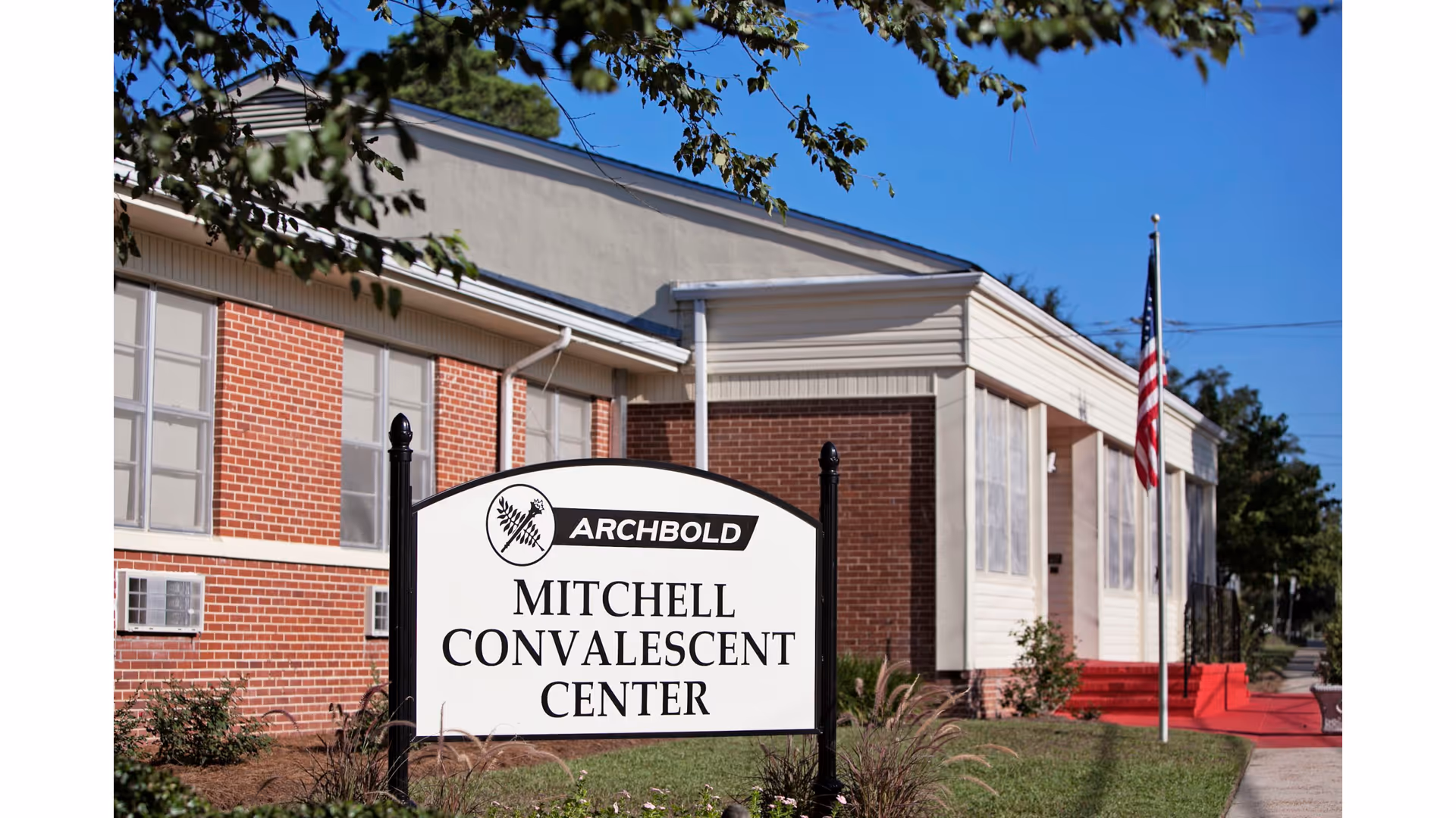 Exterior view of the Mitchell Convalescent Center building with a sign in front that reads 'Archbold Mitchell Convalescent Center'. The building has red brick and white siding, with an American flag on a flagpole near the entrance and a clear blue sky overhead.