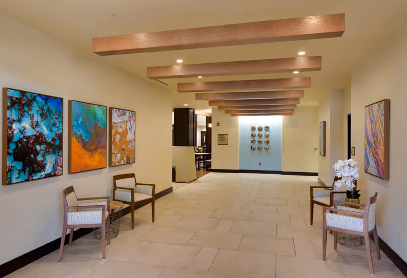 A spacious hallway in a senior living facility with beige tiled floors and cream-colored walls. The ceiling features wooden beams with recessed lighting. On the left wall, there are three colorful abstract paintings above two wooden chairs with white cushions. On the right side, there are two similar chairs with a small table holding a white orchid plant. At the end of the hallway, a blue accent wall displays a decorative arrangement of circular art pieces.