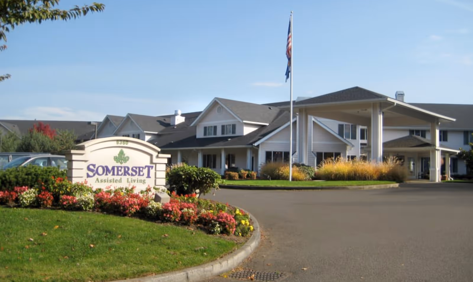 Front entrance of Somerset Assisted Living with its sign, landscaped flowerbeds, flagpole, and a covered porte-cochere.