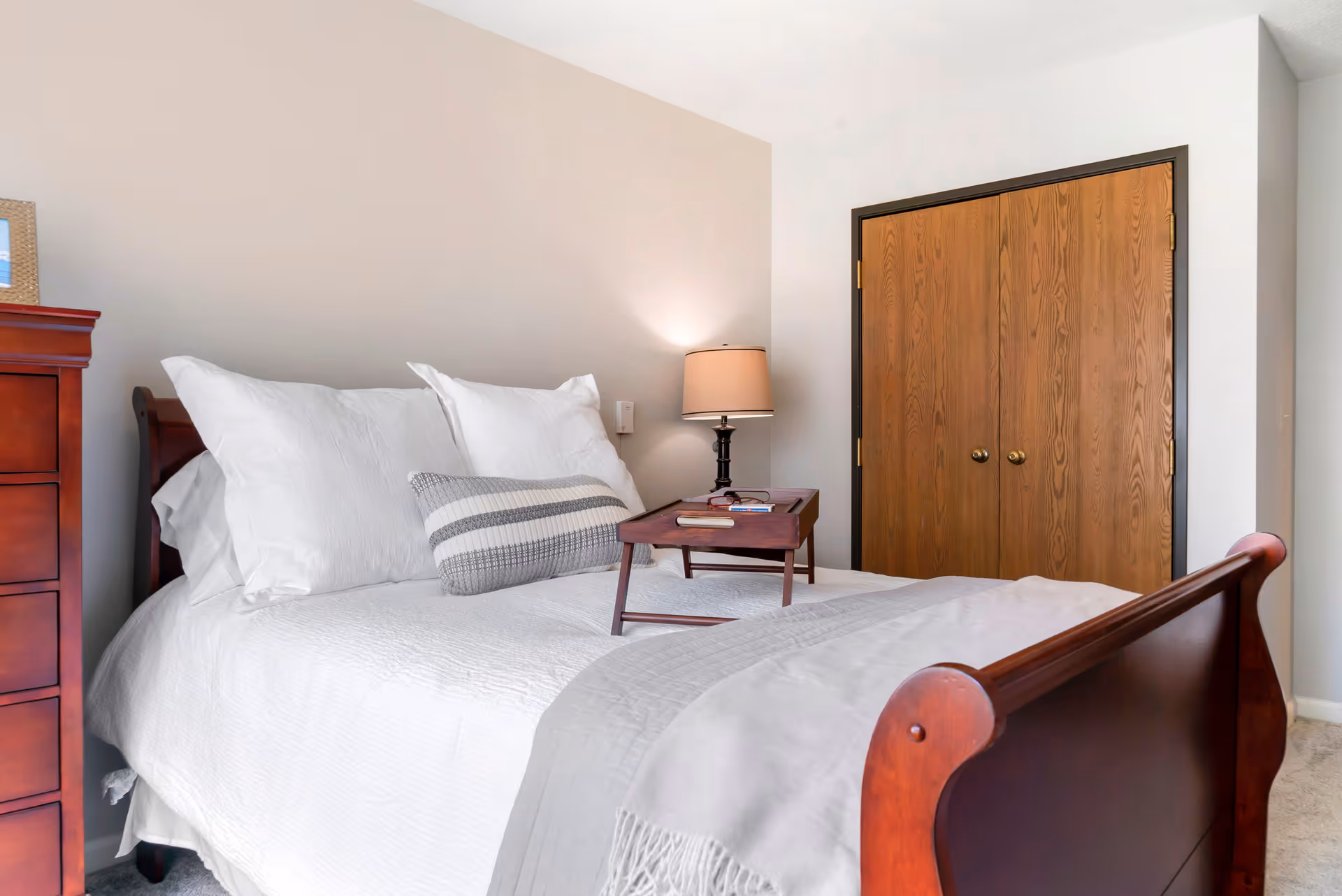 A neatly made bed with white bedding and multiple pillows in a bedroom. There is a wooden tray with books and a lamp on the bed. To the right, there is a wooden double-door closet, and to the left, a wooden dresser is partially visible.