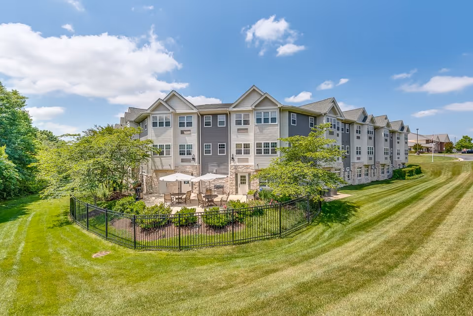 Three-story senior living building with a fenced patio area, umbrellas, and a wide manicured lawn under a blue sky.