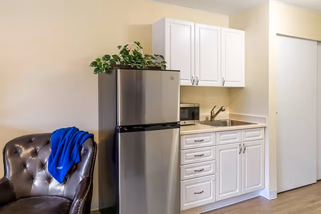 A small kitchenette area with white cabinets, a stainless steel refrigerator, a microwave, and a sink. To the left of the kitchenette is a dark brown leather chair with a blue blanket draped over the back. The walls are light-colored and the floor is wood.