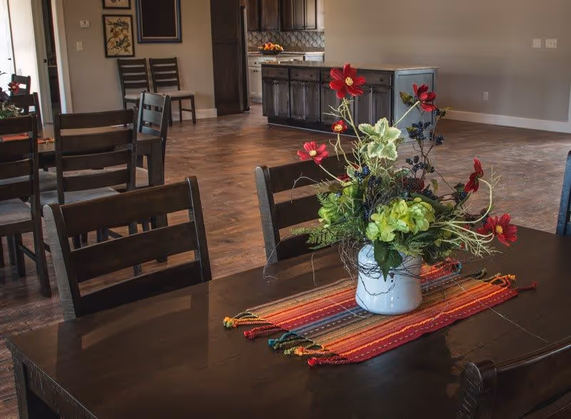 A dining area with a dark wooden table and chairs. On the table is a colorful striped runner and a white vase with a floral arrangement featuring red and green flowers. In the background, there is a kitchen island with dark cabinetry and a tiled backsplash.