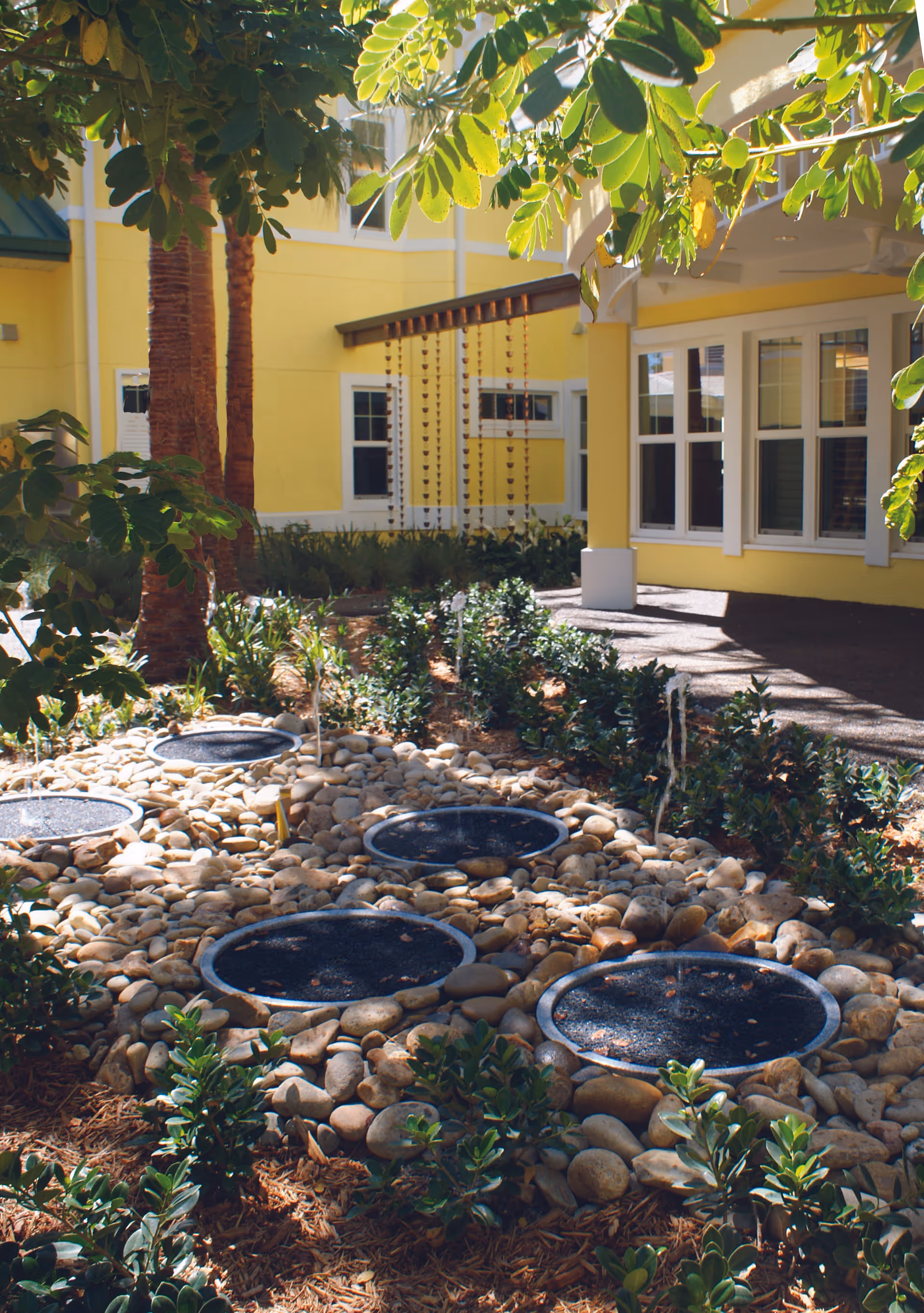 Outdoor garden area with small fountains surrounded by rocks and green plants, shaded by tree leaves, adjacent to a yellow building with white-framed windows.