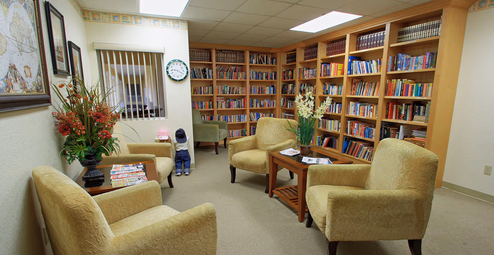 A cozy library room in an assisted living facility with four beige upholstered armchairs arranged around two wooden coffee tables. The tables have magazines and a vase with white flowers. The back wall is lined with wooden bookshelves filled with books. There is a green armchair in the corner and a large floral arrangement on a side table. A clock is mounted on the wall above a window with vertical blinds, and two small child mannequins wearing hats are placed near the wall.