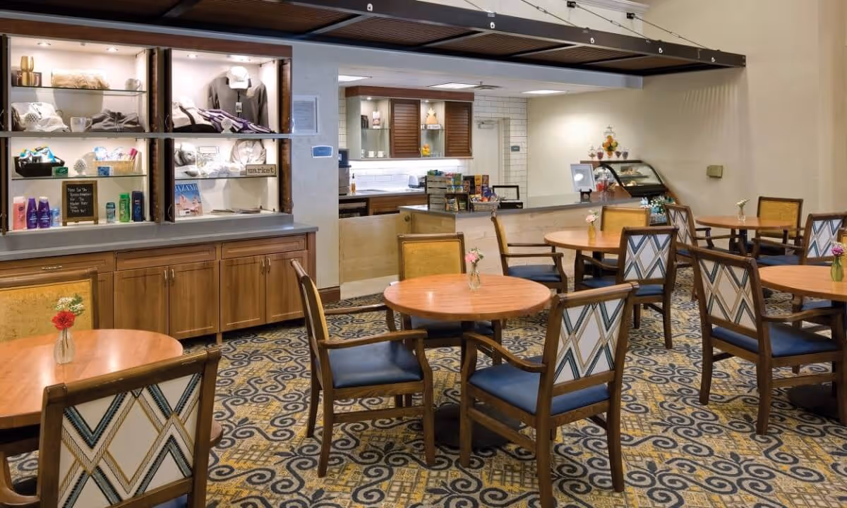 Dining room with round wooden tables and patterned chairs, a service counter and display shelves in the background.