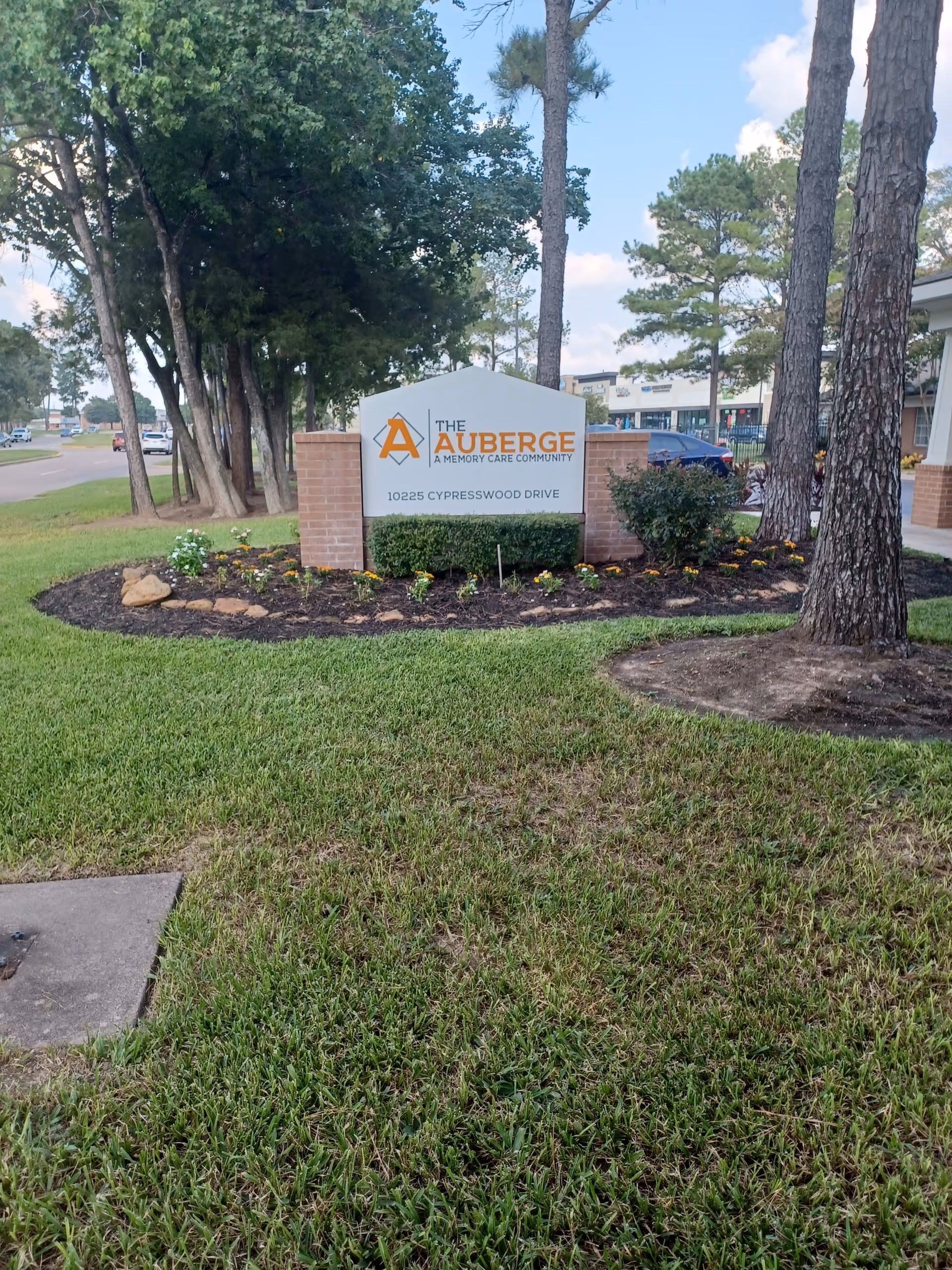Outdoor view of a landscaped area with a sign for The Auberge, a memory care community, located at 10225 Cypresswood Drive. The sign is surrounded by flowers and bushes, with trees and a road visible in the background.