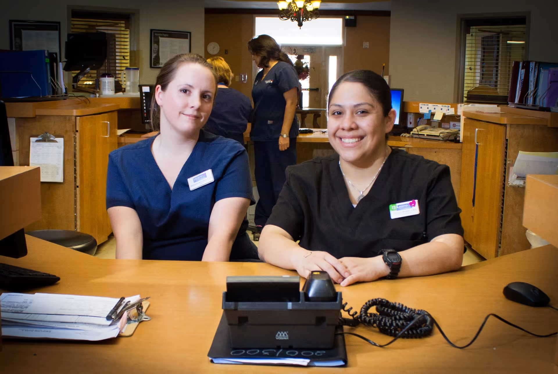 Two healthcare workers sitting behind a reception desk in a healthcare facility. Both are wearing scrubs and name badges, smiling at the camera. In the background, two other healthcare workers are engaged in conversation near a doorway.
