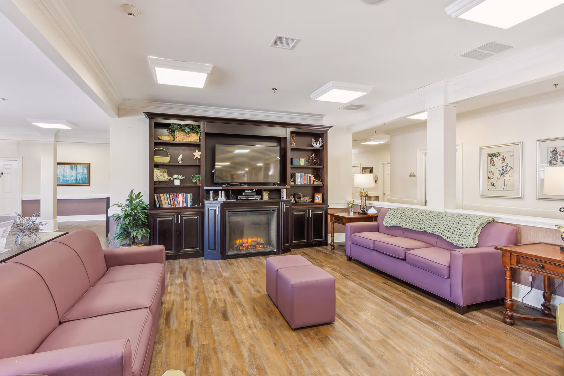 A cozy living room area with two purple sofas facing a dark wooden entertainment center that houses a flat-screen TV and an electric fireplace. The room has wooden flooring, a small purple ottoman in the center, side tables with lamps, and framed floral artwork on the walls.