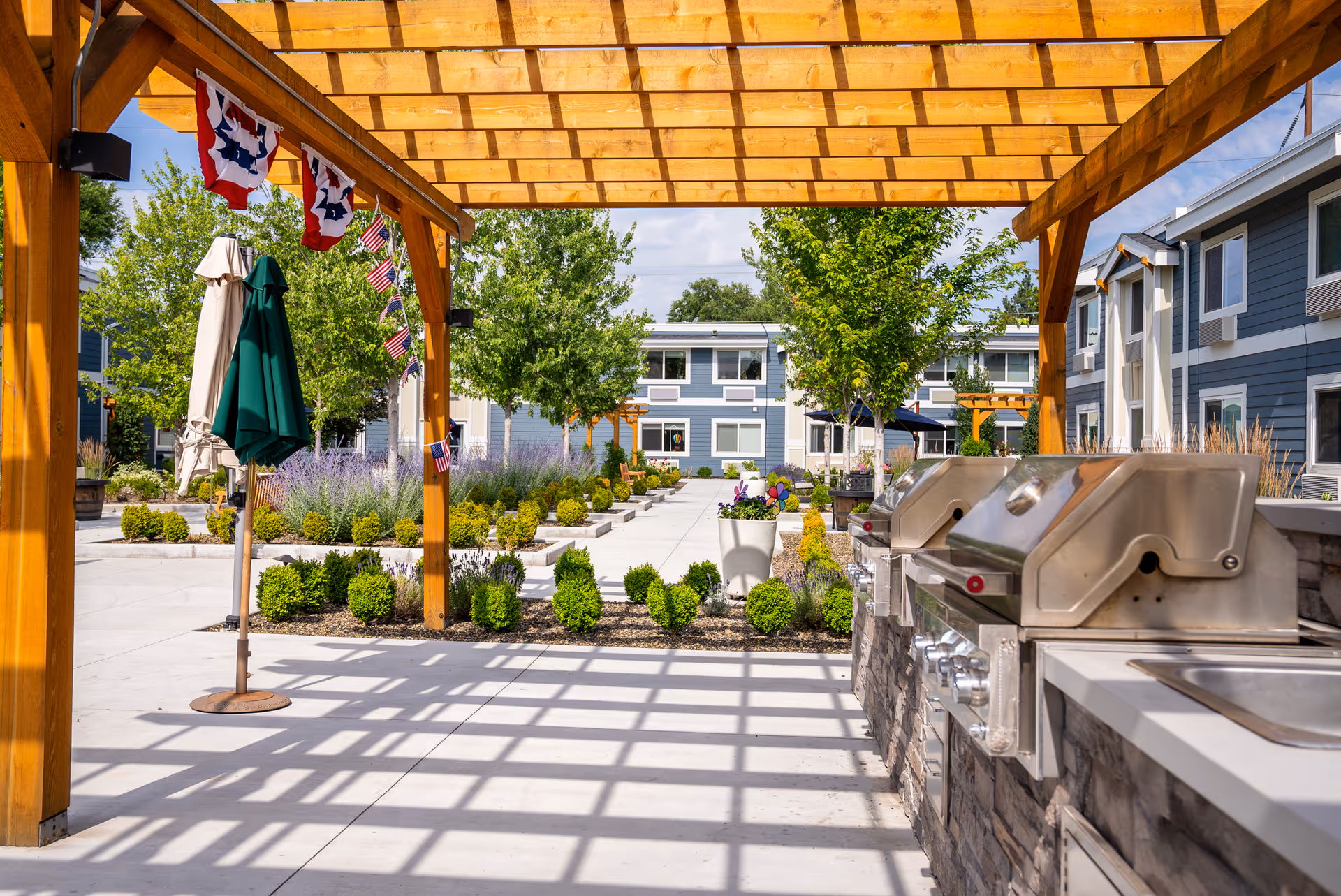 Outdoor patio area at Heatherwood Senior Living featuring a wooden pergola casting shadows on the concrete floor, two closed umbrellas, stainless steel grills built into a stone counter, and landscaped garden beds with bushes and trees. Residential buildings are visible in the background under a partly cloudy sky.