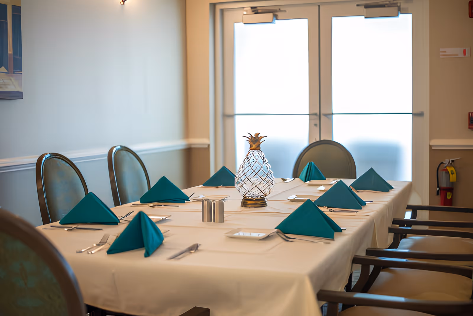 A dining table set for a meal with teal folded napkins, silverware, small plates, and a decorative glass pineapple centerpiece in a well-lit room with chairs around the table and double doors in the background.