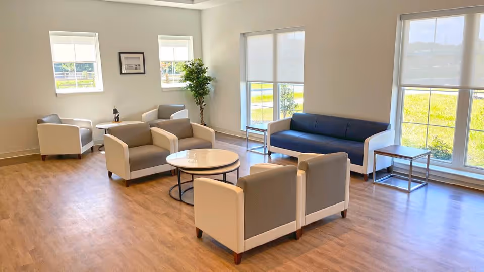 A bright and spacious senior living facility common area with large windows letting in natural light. The room features a blue sofa, several gray and white armchairs arranged around two round coffee tables, a potted plant in the corner, and framed artwork on the wall. The floor is wood, and the overall atmosphere is clean and inviting.