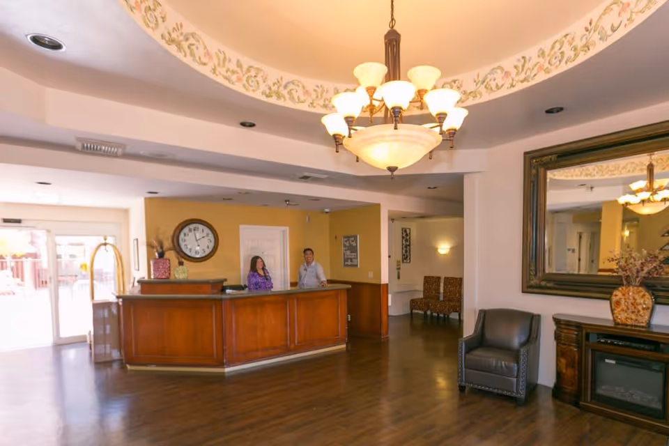 Reception area of Lexington Assisted Living with a wooden front desk where two staff members stand behind. The room features a large decorative chandelier, a wall clock, a large mirror above a wooden cabinet with a vase, and seating area with chairs. The floor is wooden and the walls are painted in warm tones.