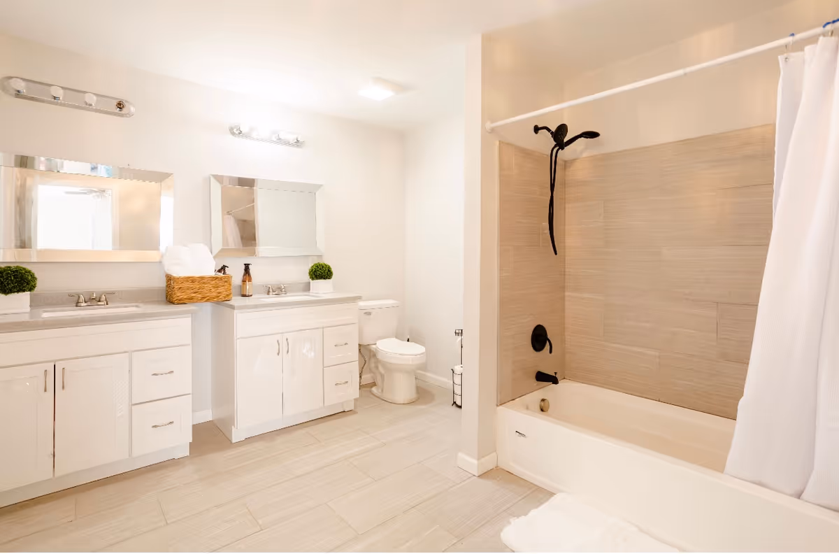 A bright and clean bathroom featuring a double sink vanity with two mirrors above, a toilet, and a bathtub with a showerhead and a white shower curtain. The floor is tiled, and the walls are painted white. There are small green plants on the vanity and a basket with towels.