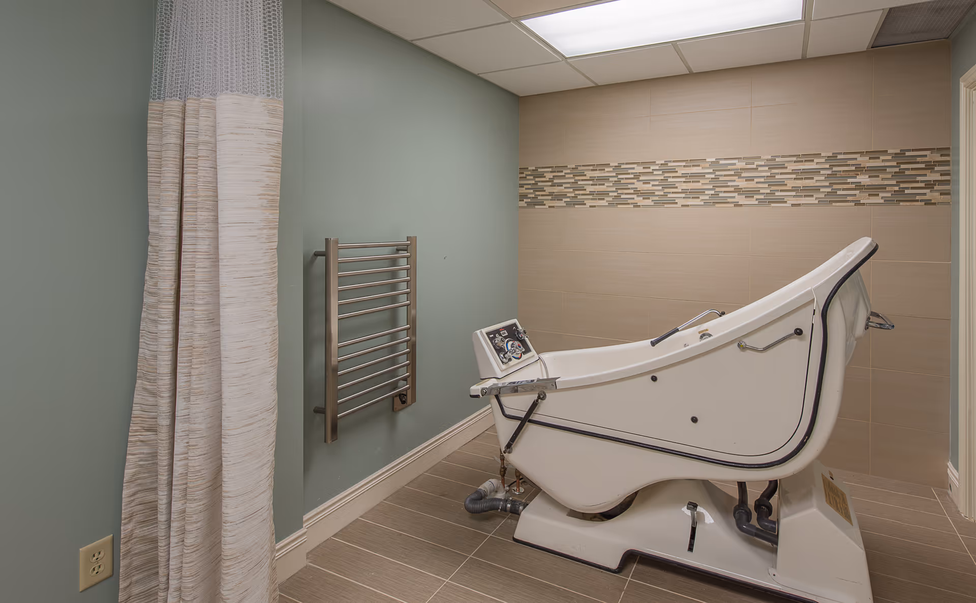 A reclining assisted bathing tub in a tiled room with a privacy shower curtain and a heated towel rack.