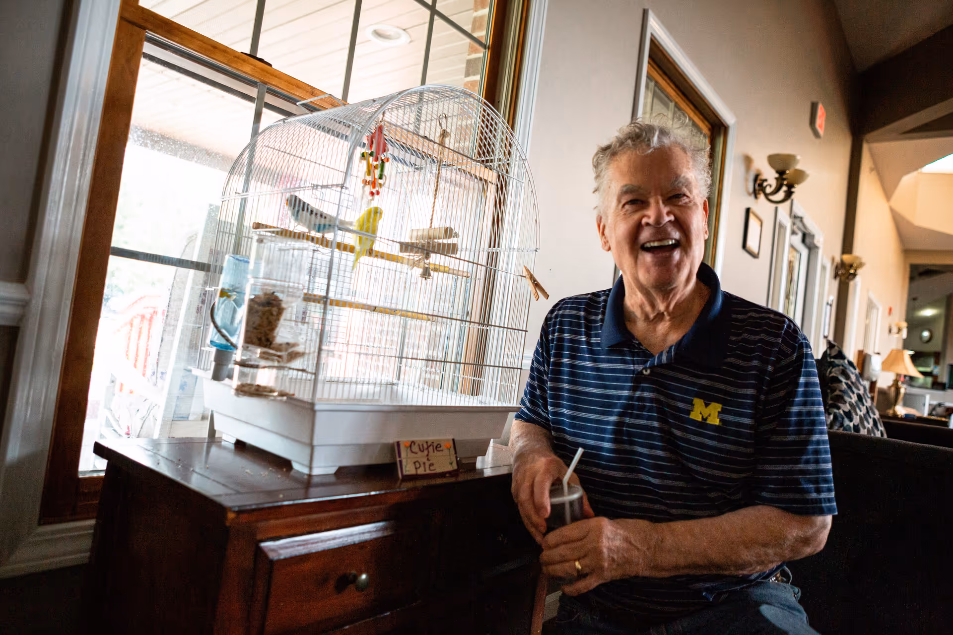 An elderly man wearing a striped polo shirt with a University of Michigan logo is sitting indoors next to a birdcage with two birds inside. The birdcage is placed on a wooden cabinet near a large window. The man is smiling and holding a drink with a straw. The room has wall sconces and framed pictures on the wall.