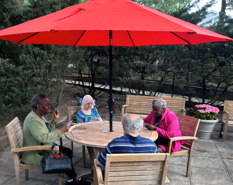 Four older adults sitting around a wooden outdoor table under a large red patio umbrella.