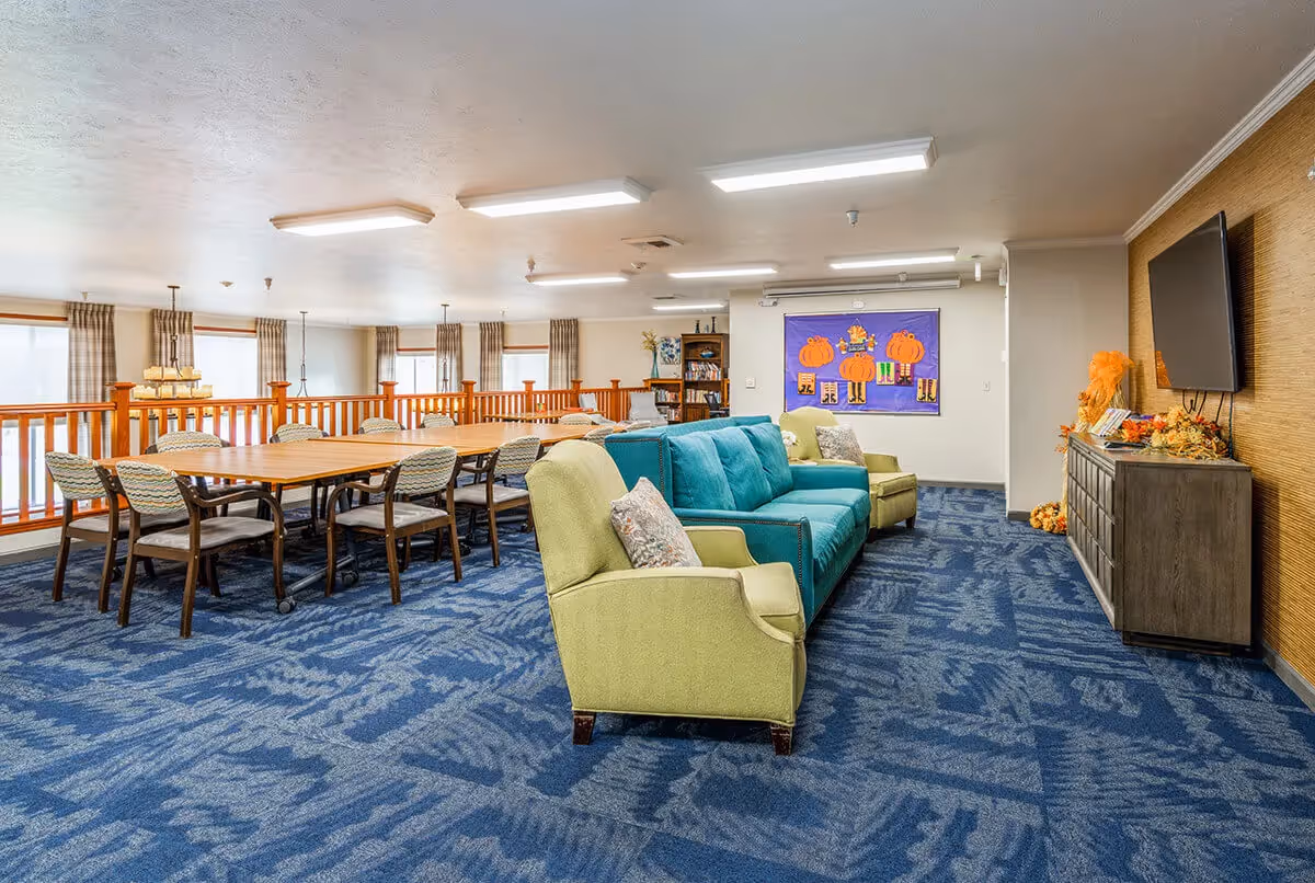 A spacious common area with a blue patterned carpet, featuring a teal sofa and two green armchairs with decorative pillows. Behind the seating area is a long wooden table with multiple chairs. The walls are decorated with a colorful bulletin board with pumpkin-themed artwork and a large flat-screen TV mounted above a wooden cabinet adorned with fall decorations. Large windows with plaid curtains allow natural light into the room.