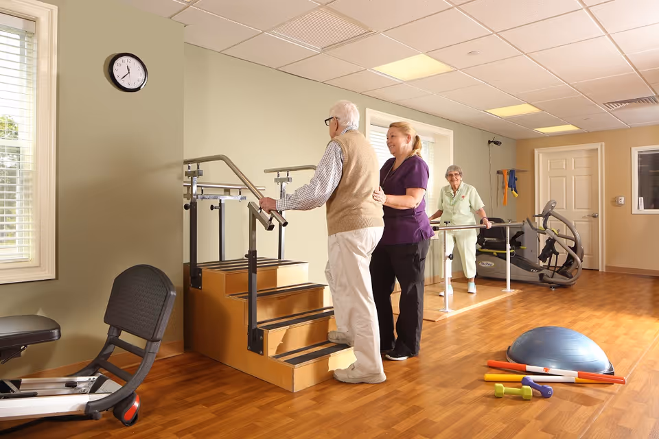An elderly man is assisted by a caregiver as he practices walking up a small set of stairs with handrails in a rehabilitation or physical therapy room. Another elderly woman is using parallel bars for walking exercises in the background. The room has wooden flooring, exercise equipment, and large windows letting in natural light.
