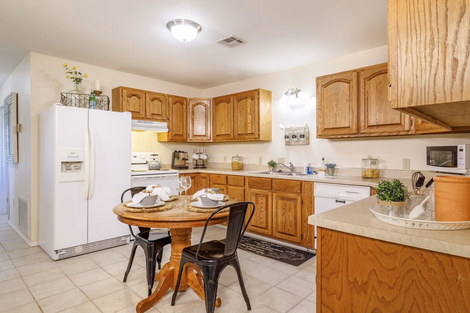 A bright kitchen with wooden cabinets, a white refrigerator, stove, and dishwasher. There is a small round wooden table set for two with plates, bowls, napkins, and wine glasses. The countertop has various kitchen items including a coffee maker, a jar of pasta, a small plant, and a microwave. The floor is tiled and the walls are light-colored.