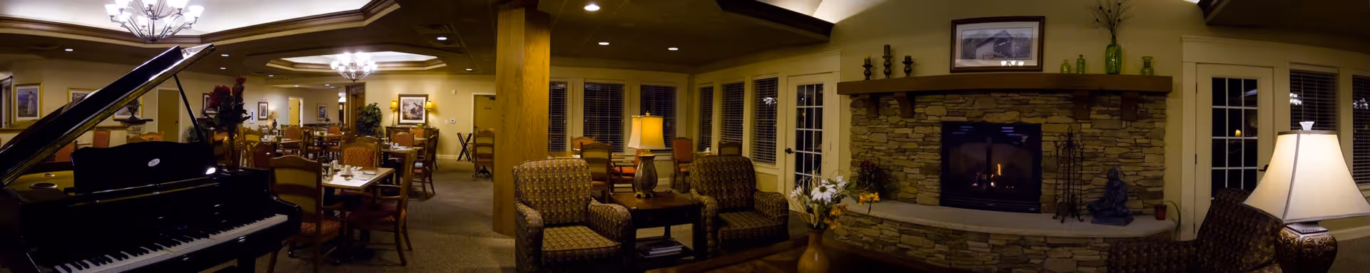 Interior view of a senior living facility common area featuring a grand piano on the left, multiple dining tables and chairs in the center and background, two upholstered armchairs with a side table and lamp in the foreground, and a stone fireplace with decorative items on the right. The room is warmly lit with ceiling lights and table lamps, and has large windows with blinds.
