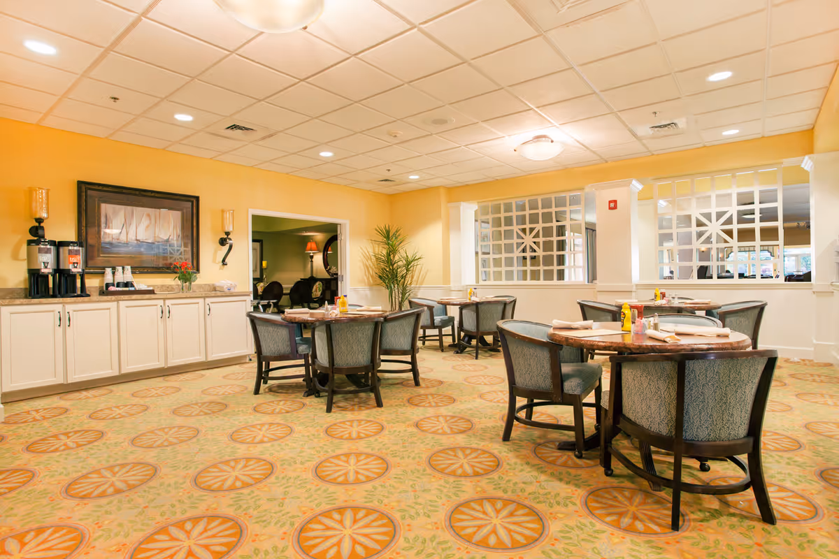 A dining room with round tables and cushioned chairs arranged neatly. The room has a patterned carpet with orange and green designs, yellow walls, and a white ceiling with recessed lighting. On the left side, there is a counter with coffee dispensers, cups, and a framed painting of sailboats above it. A plant is placed near the entrance to an adjoining room.