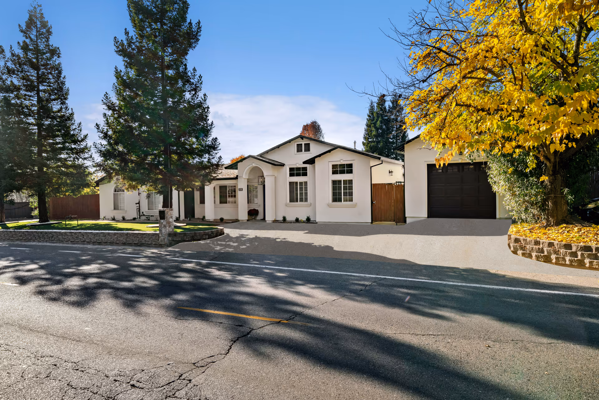Exterior view of a single-story white building with a black garage door, surrounded by tall evergreen trees and a tree with yellow autumn leaves, under a clear blue sky.