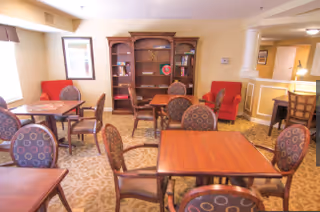 A cozy common area with several wooden tables and cushioned chairs arranged on a patterned carpet. There is a large wooden bookshelf against the back wall, a red armchair, and a window letting in natural light. The walls are painted a warm beige color with white trim.
