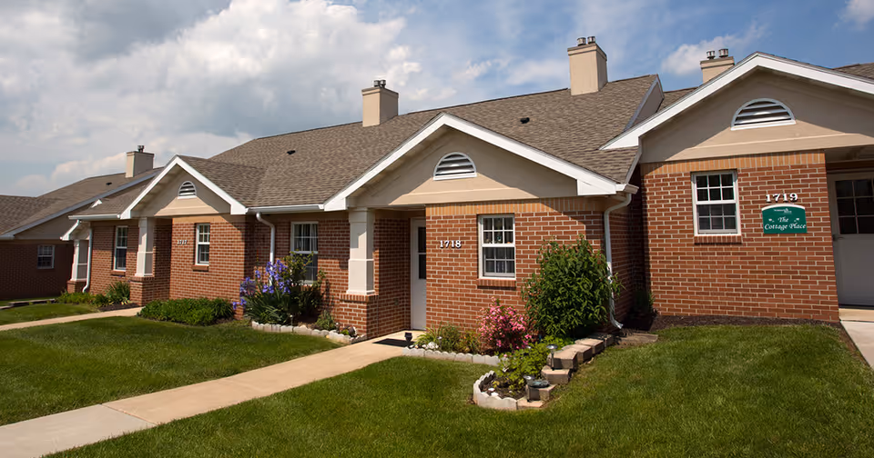 Exterior view of a single-story brick building with multiple entrances, each with a small garden and walkway leading up to the doors. The building has a brown shingled roof with chimneys and white trim around the windows and doors. The sky is partly cloudy.