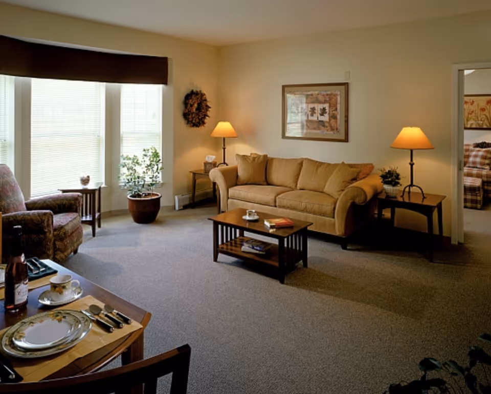 Well-lit living room with a sofa, armchair, coffee table, side tables with lamps, dining place setting, and large windows.