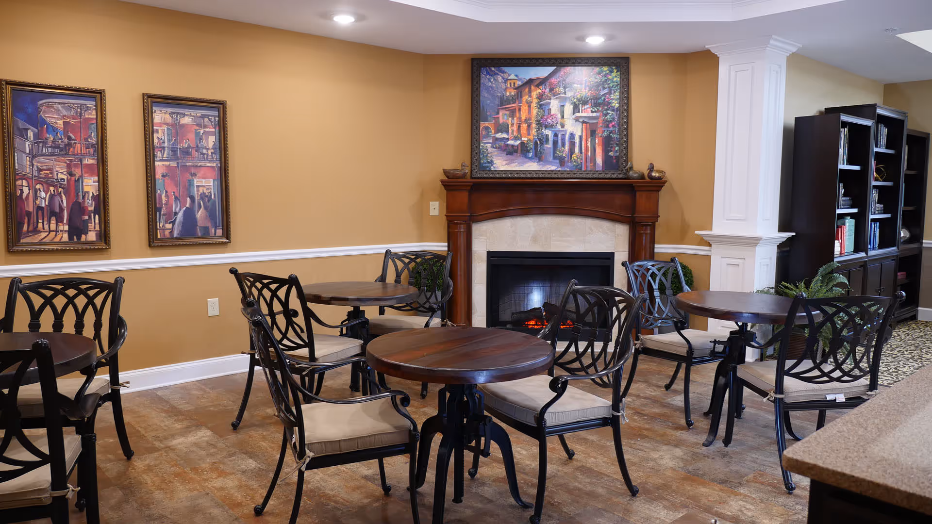 A cozy common area with several round wooden tables and black metal chairs with beige cushions. The room features a fireplace with a wooden mantel, above which hangs a colorful painting of a street scene. Two framed paintings depicting people are on the left wall, and a dark wooden bookshelf is visible on the right side. The walls are painted a warm beige color, and the floor has a rustic tile appearance.