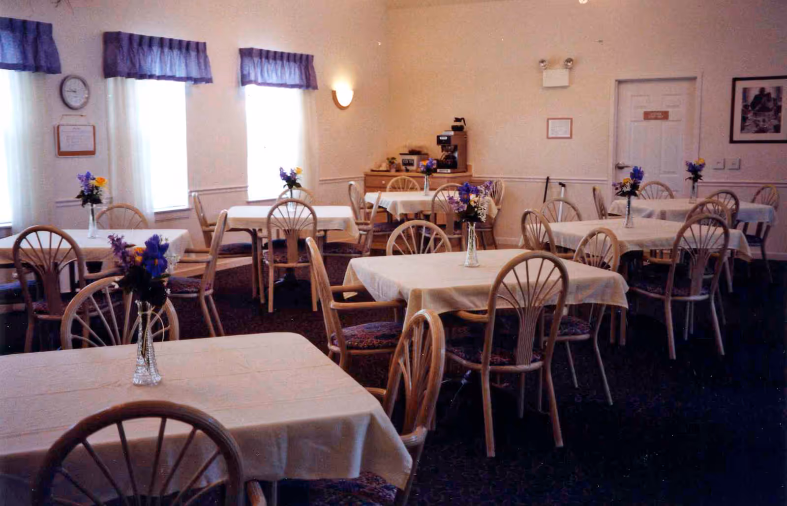 A dining room with multiple tables covered in white tablecloths, each table decorated with a small vase of purple and yellow flowers. Wooden chairs with patterned cushions surround the tables. The room has three windows with blue valances, a clock on the wall, and a sideboard with a coffee machine and additional flowers.