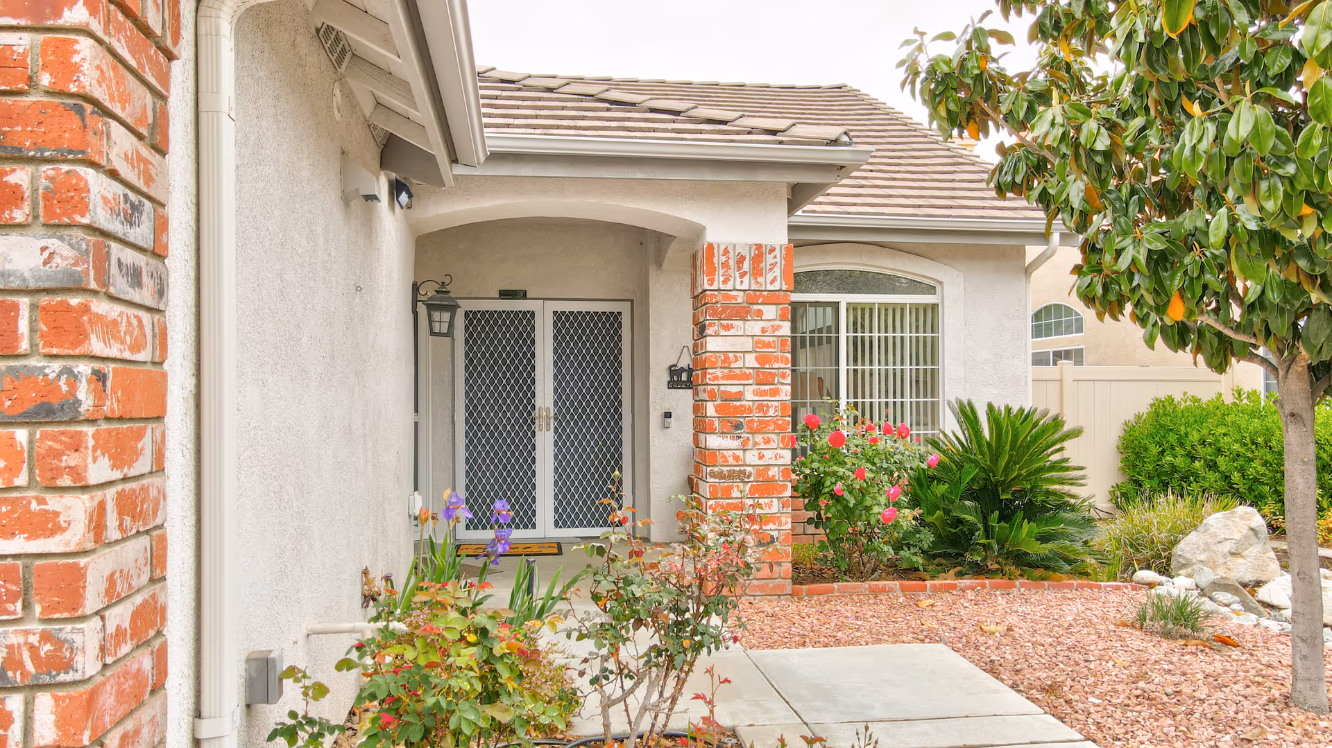 Front entrance of a single-story home with a covered porch, white double doors with security screens, brick columns, and a landscaped front yard featuring flowering plants, bushes, a tree, and decorative rocks.