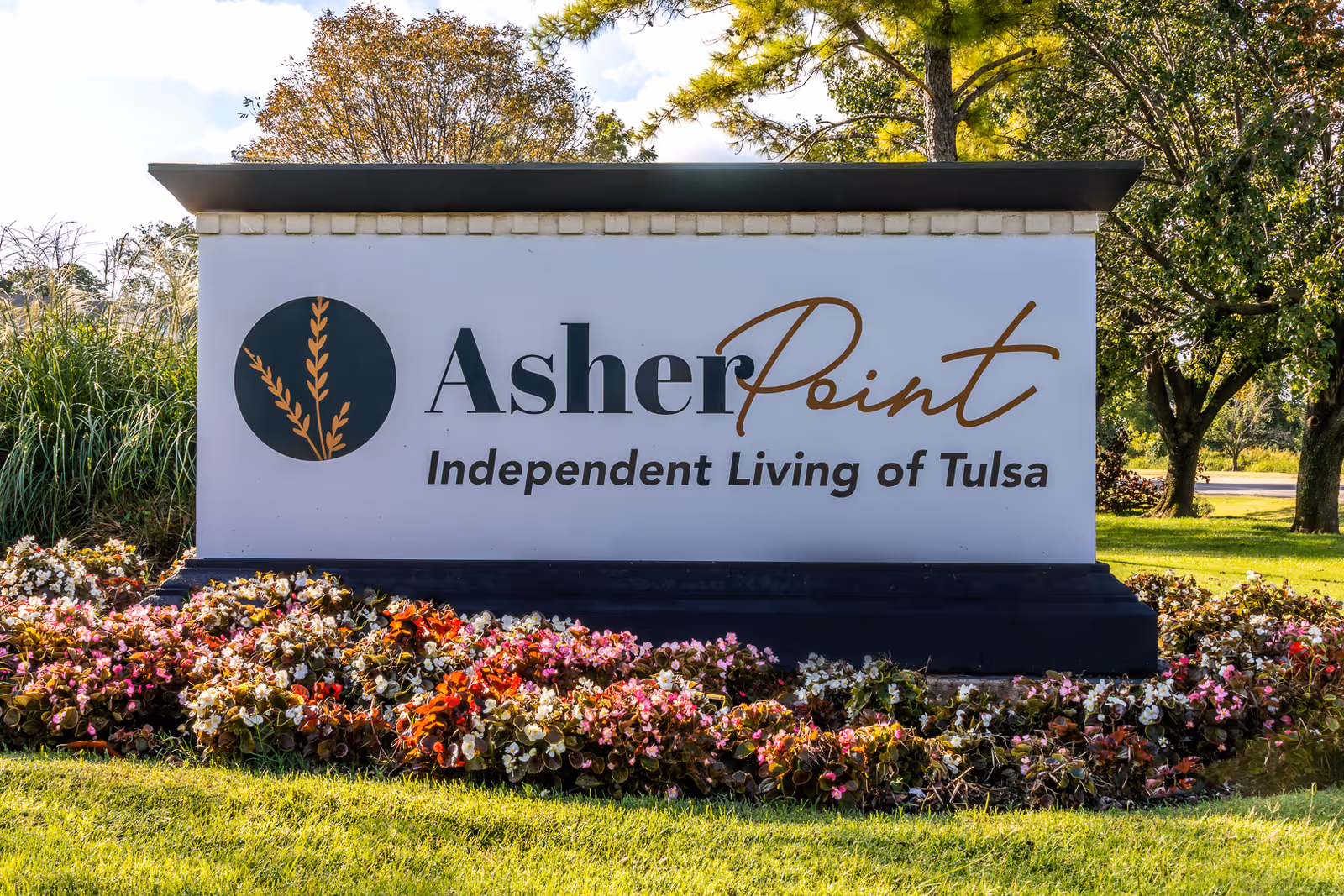 A large white sign with the logo and text 'Asher Point Independent Living of Tulsa' surrounded by colorful flowers and green grass, with trees and a blue sky in the background.