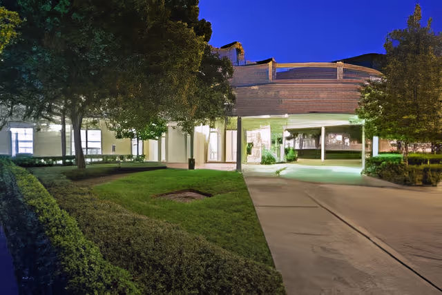 Exterior view of a senior living facility at night with well-lit entrance, surrounded by trees, bushes, and a neatly maintained lawn under a clear dark blue sky.
