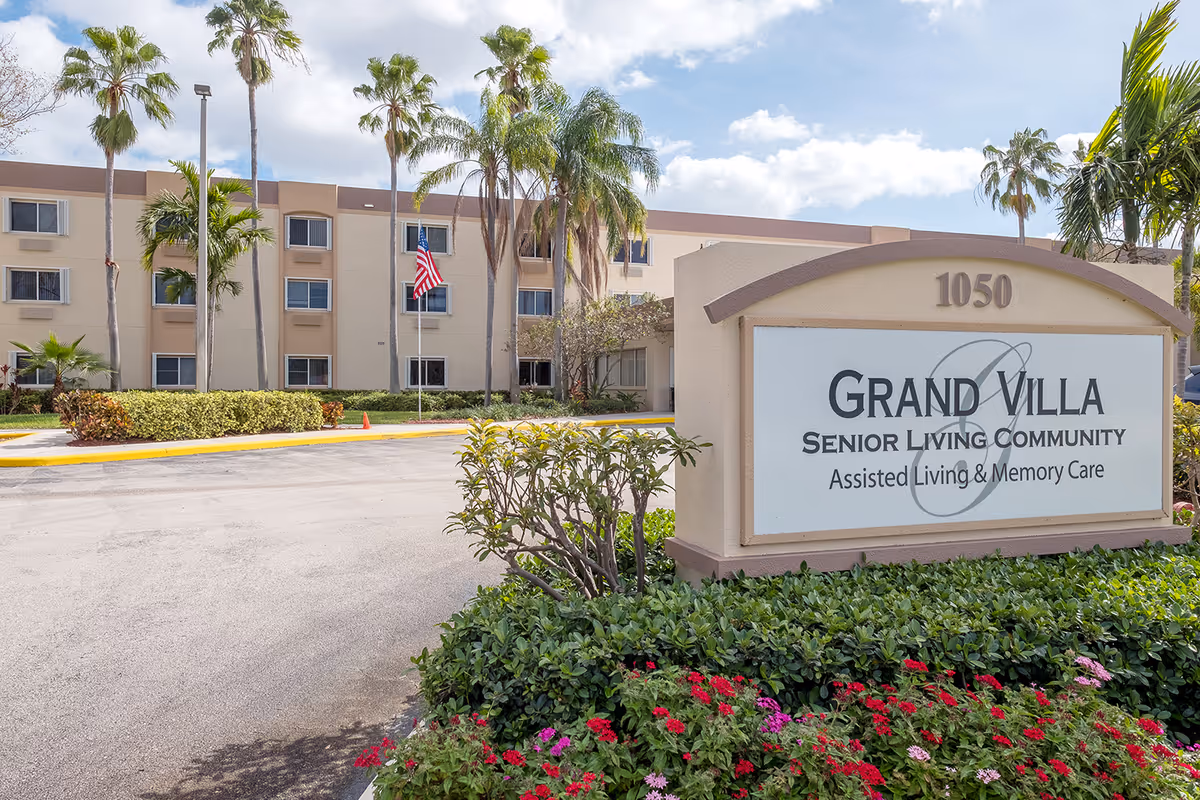 Entrance sign and front facade of the Grand Villa senior living community with palm trees and landscaped grounds.