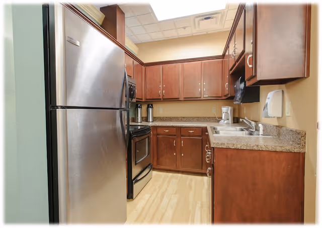 A small kitchen area with wooden cabinets, a stainless steel refrigerator, an oven with a stove, a coffee maker, and a double sink. The countertops are granite, and the floor is light wood. The walls are painted beige, and there is a ceiling light panel.