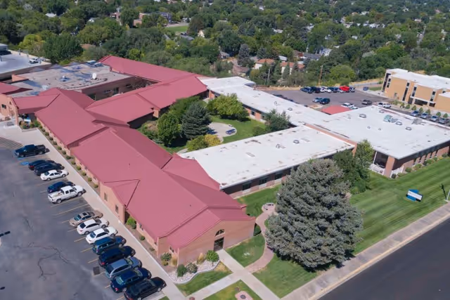 Aerial view of Gateway Transitional Care Center showing a large building complex with red and white roofs surrounded by green trees and lawns. There is a parking lot with several cars parked along one side of the building and a road adjacent to the property.