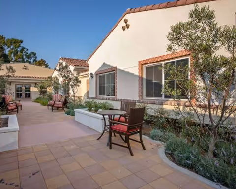 Sunny courtyard with tiled patio, outdoor tables and chairs, planter beds and a single-story Mediterranean-style building.