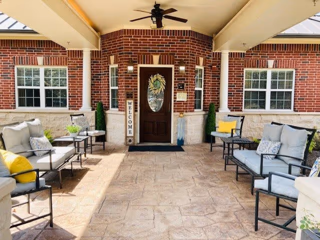 Covered outdoor patio area with cushioned seating including sofas and chairs arranged along both sides. The space features a ceiling fan, brick walls, windows with white shutters, and a dark wooden door with a decorative glass panel. A vertical 'WELCOME' sign is placed next to the door, and there are small plants and side tables near the seating.